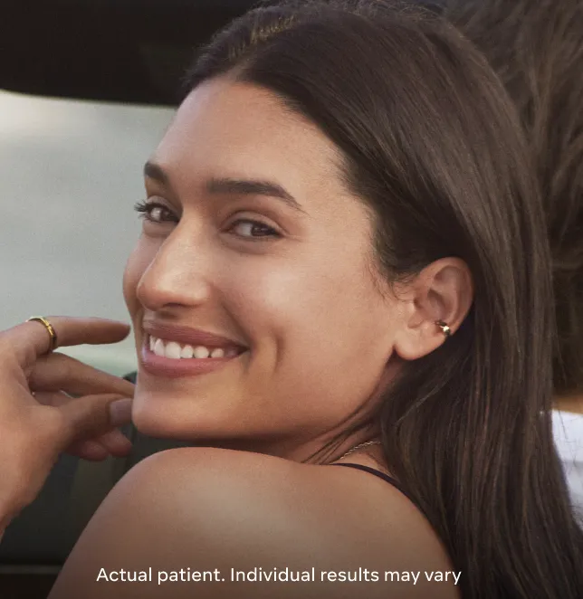 Smiling young woman with long dark hair, wearing a gold ring and earrings, looks over her shoulder. The image conveys a sense of confidence and satisfaction, showcasing an actual patient. The background suggests a casual outdoor setting, emphasizing a natural and relatable appearance. This image highlights the potential results of cosmetic procedures, appealing to those considering similar treatments. Individual results may vary, as noted.