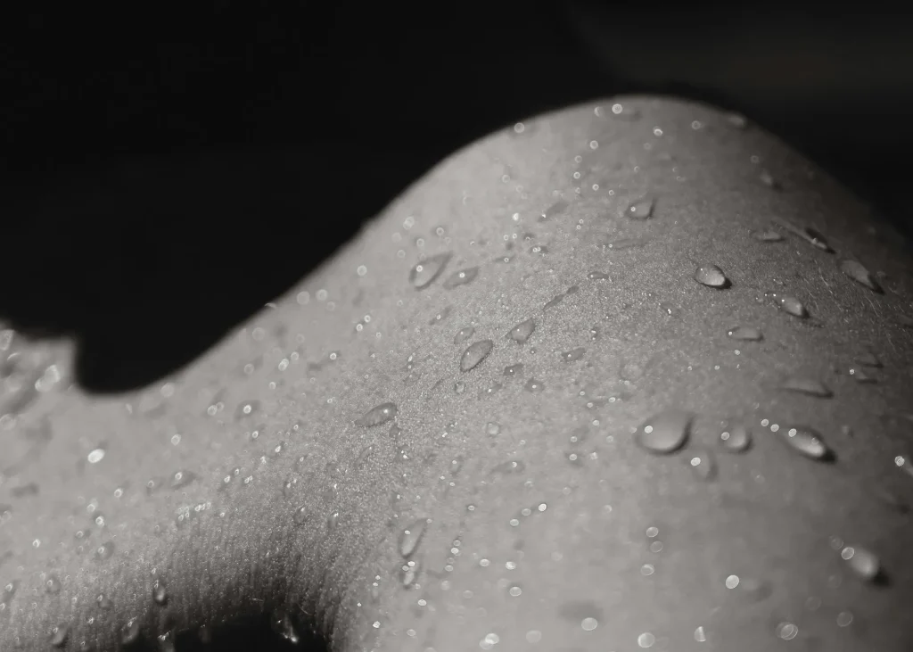 Close-up of a textured surface with water droplets glistening in black and white. The image captures the details of the skin's surface, highlighting the contrast between light and shadow. The droplets vary in size, creating a visually appealing pattern that emphasizes the smoothness of the skin beneath. This artistic representation showcases the natural beauty of water on skin, evoking a sense of freshness and vitality. Ideal for topics related to skincare, hydration, or artistic photography.