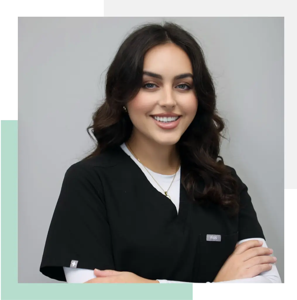Smiling female healthcare professional in a black uniform with a name tag, standing confidently with arms crossed. The background features a modern design in soft colors. This image represents professionalism and approachability in the healthcare field, ideal for showcasing staff or services on a medical website or promotional material.