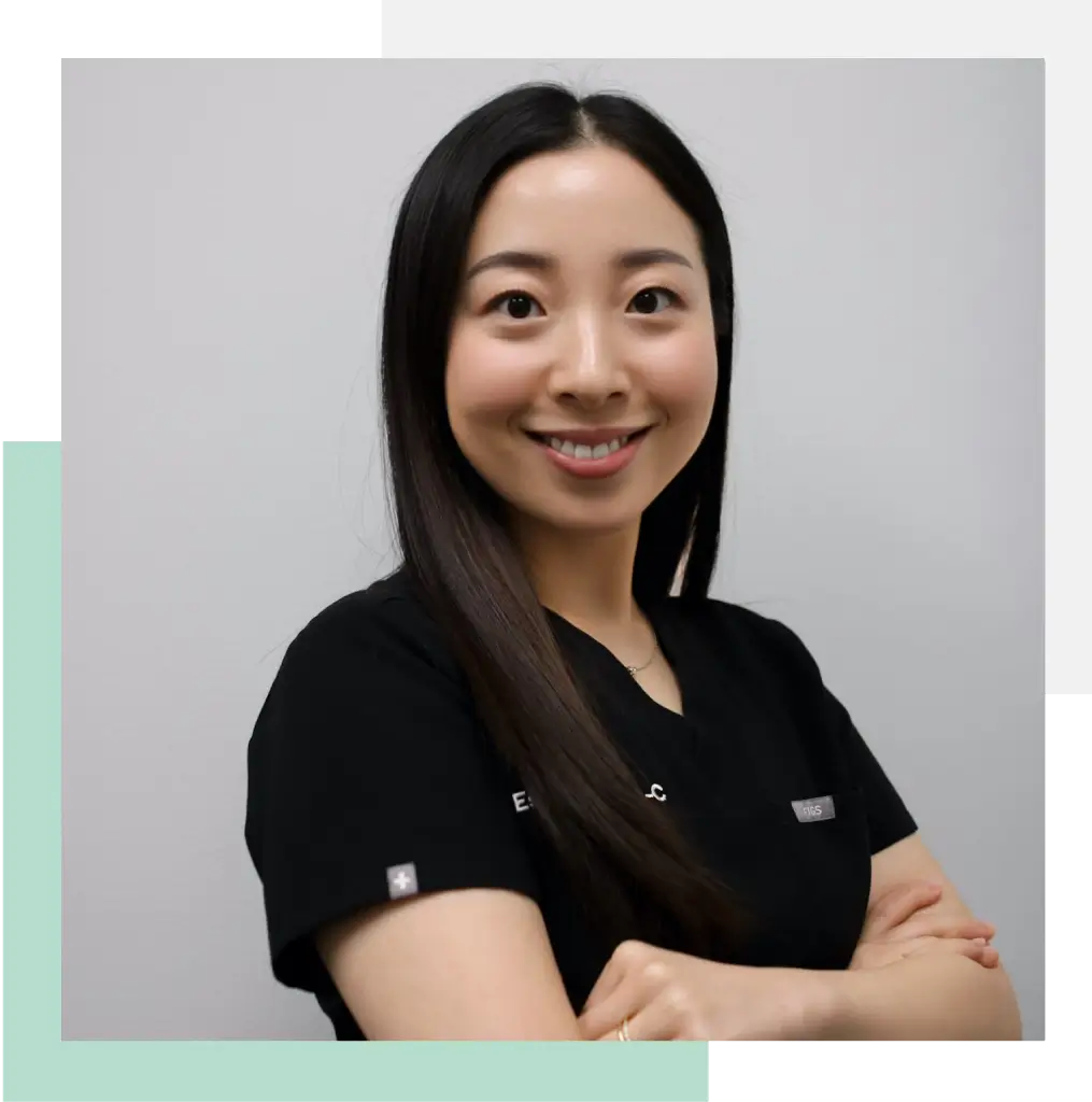 Professional portrait of a smiling woman wearing a black medical uniform, with her arms crossed. She has long dark hair and is set against a neutral gray background. This image represents healthcare professionals dedicated to patient care, emphasizing approachability and expertise in a clinical environment. Ideal for showcasing medical services or staff on a healthcare website or promotional materials.