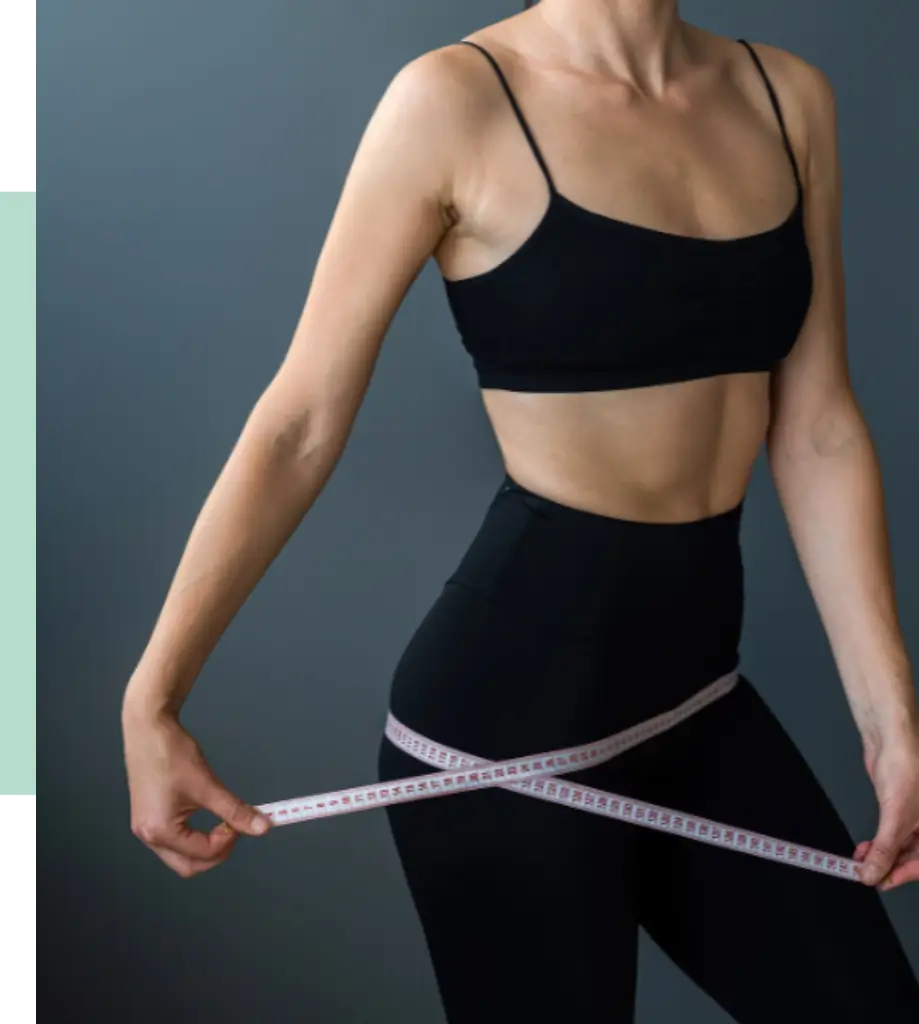 A woman in a black crop top and high-waisted leggings measures her waist with a tape measure. The background is a neutral gray, emphasizing her athletic physique. This image represents themes of fitness, body measurement, and health awareness, making it suitable for articles or content related to wellness, personal training, or body positivity.
