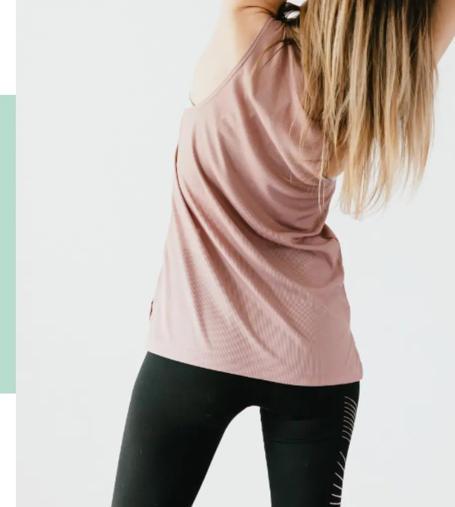 A woman in a pink athletic tank top and black leggings is seen from behind, showcasing a relaxed pose. The tank top features a loose fit, ideal for workouts and casual wear, while the leggings have a stylish design with a unique pattern. The background is minimalistic, emphasizing the outfit's contemporary look. This image highlights activewear that combines comfort and fashion, suitable for various fitness activities.