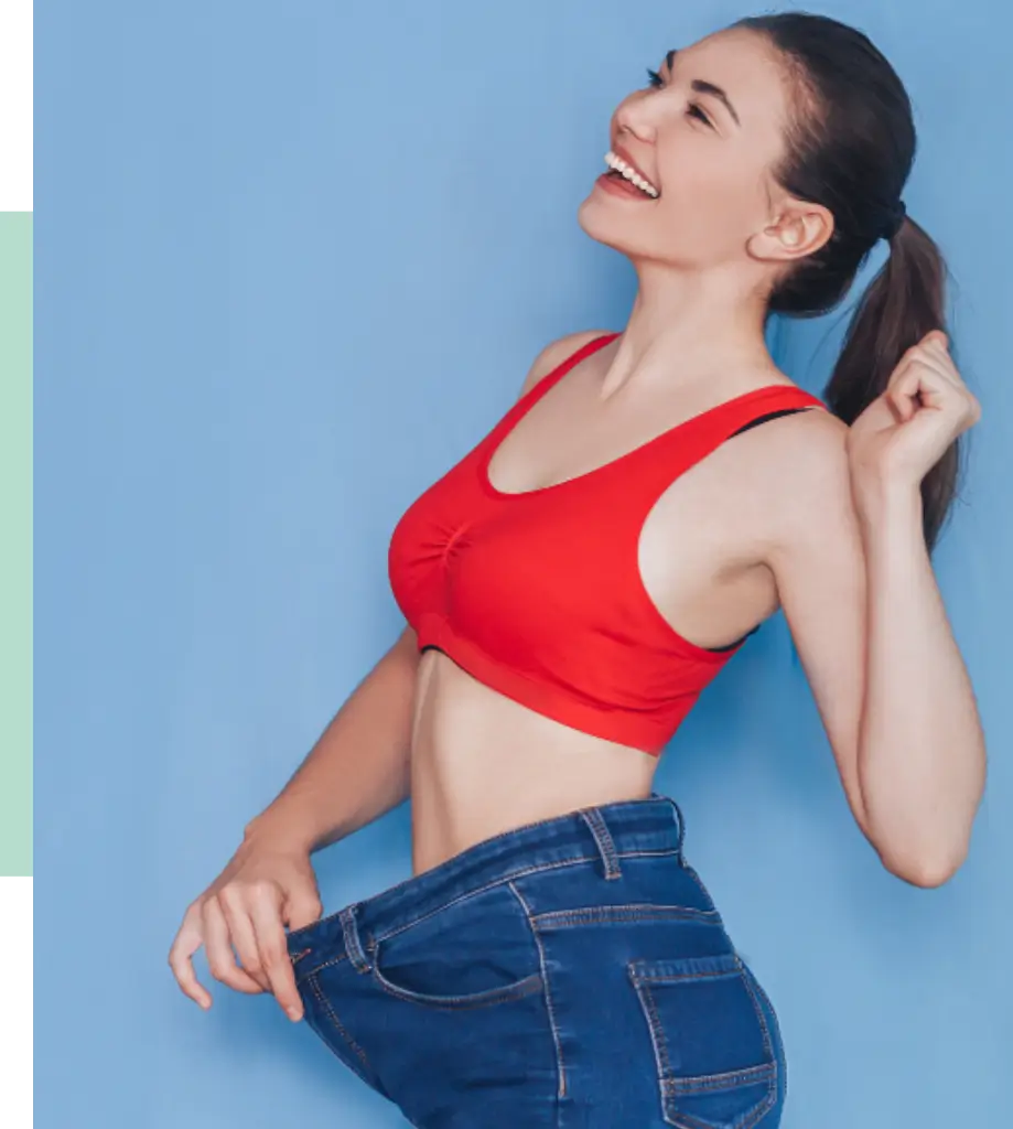 A smiling woman in a red sports bra and oversized blue jeans stands against a light blue background, celebrating a successful weight loss journey. Her joyful expression and confident posture highlight personal transformation and body positivity. This image represents fitness achievements and motivates viewers to embrace healthy lifestyle changes.