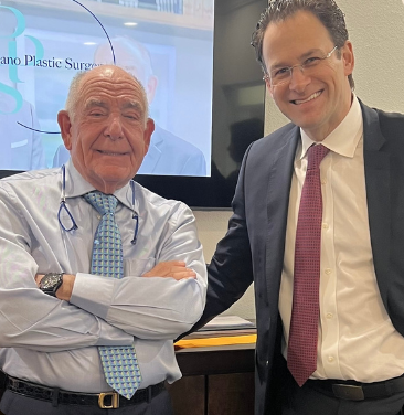 Two male plastic surgeons stand side by side, smiling confidently in a modern office setting. The older surgeon, wearing a light blue dress shirt and glasses, has his arms crossed and exudes a sense of experience. The younger surgeon, dressed in a dark suit and pink tie, stands next to him with a welcoming expression. Behind them is a digital display featuring the name of their practice, signifying a professional environment. This image represents collaboration and expertise in the field of plastic surgery, highlighting the importance of