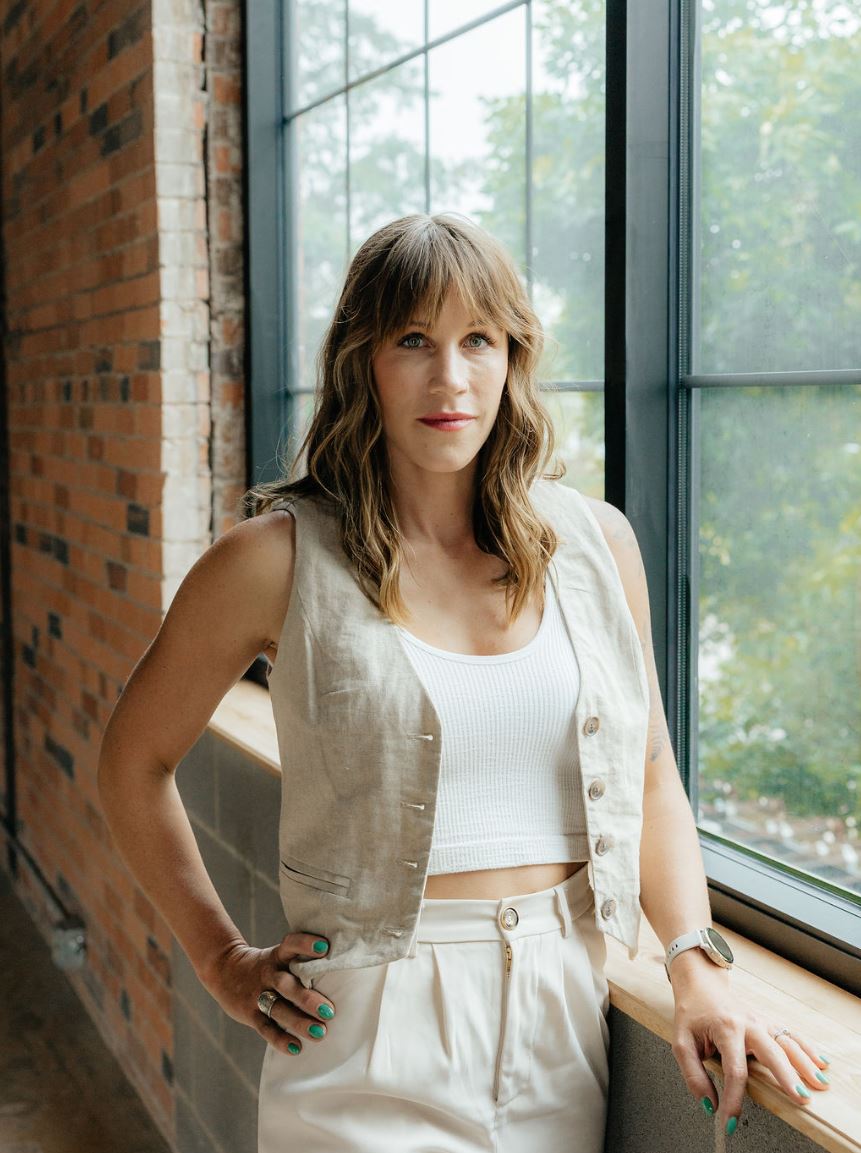 A woman with long, wavy hair stands confidently by a large window, showcasing a modern, industrial interior. She wears a light-colored linen vest over a white crop top and high-waisted cream trousers. Her left hand rests on her hip while her right hand holds a watch, and she displays vibrant green nail polish. The backdrop features exposed brick walls and greenery visible through the window, creating a bright and inviting atmosphere. This image conveys a sense of style and professionalism, perfect for lifestyle or fashion-related